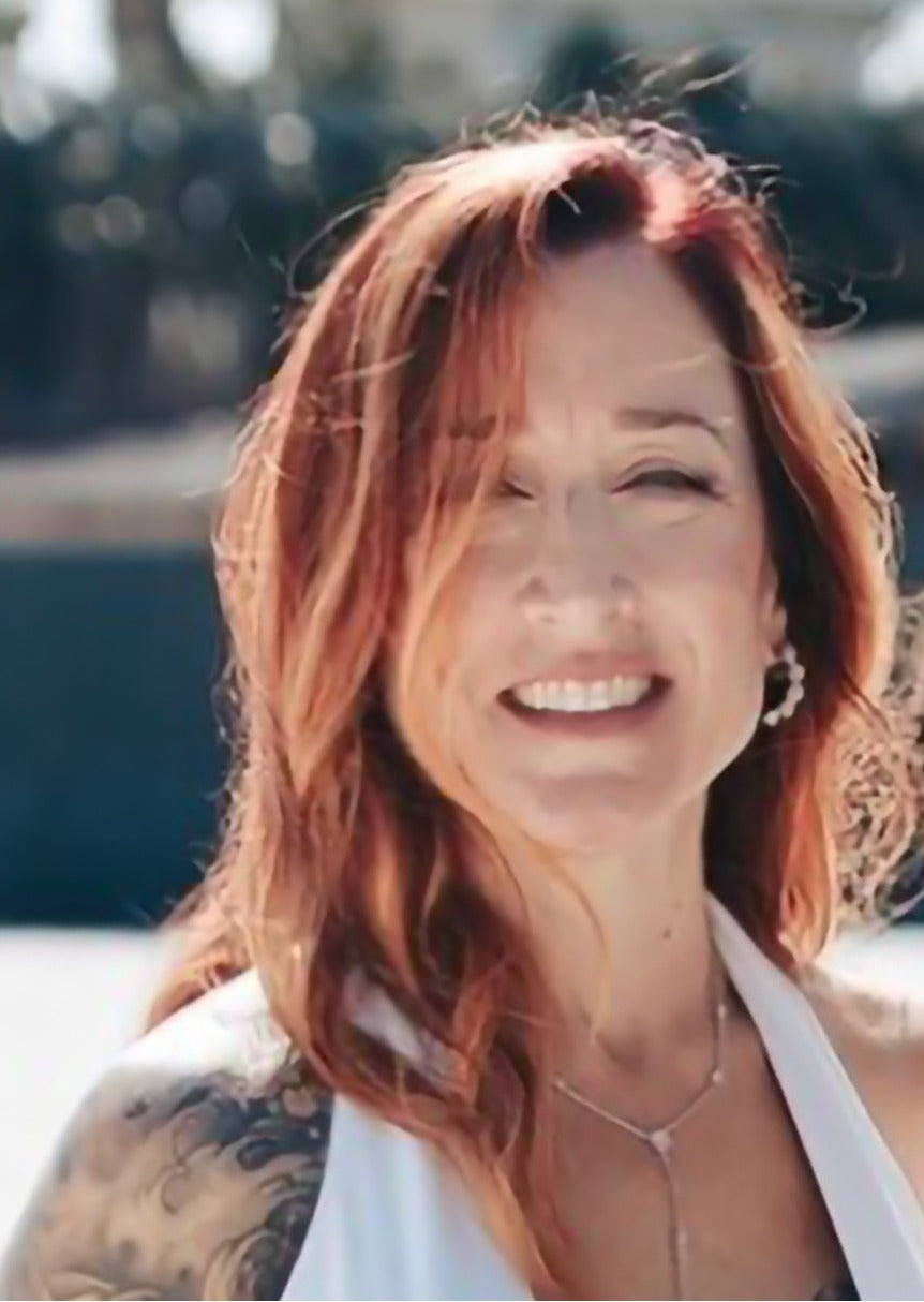 A smiling woman with long wavy red hair and a visible tattoo on her left shoulder, wearing a white halter top and a delicate silver necklace in an outdoor setting.
